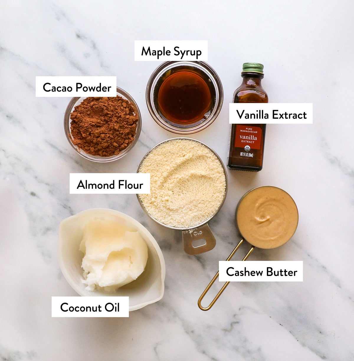 cup of coconut oil, almond flour, cashew butter, maple syrup, cacao powder, and vanilla extract on a white surface.