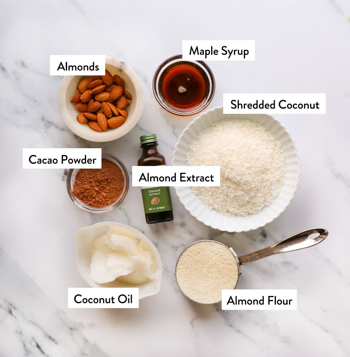 bowls of almonds, coconut, almond flour, coconut oil, and cacao powder on a white surface.