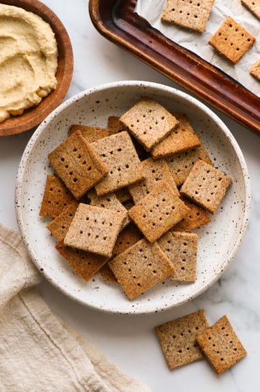 almond pulp crackers in a white bowl near hummus for serving.