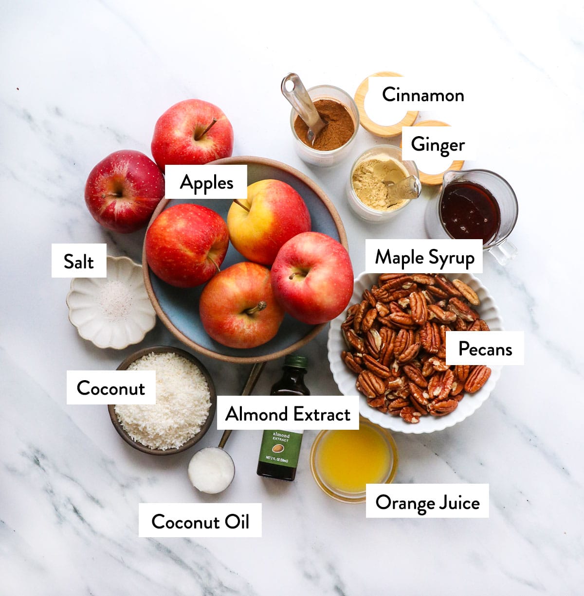 a big bowl of apples, pecans, coconut, maple syrup, and pantry spices on a white surface.
