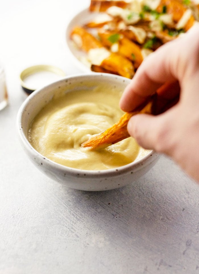 hand dipping a sweet potato fry into roasted cauliflower cheese sauce
