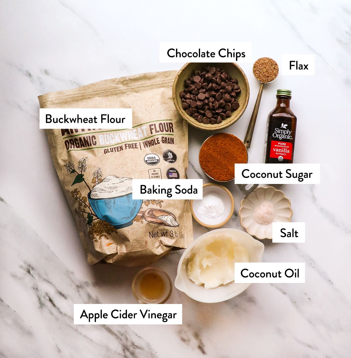 bag of buckwheat flour, bowl of chocolate chips, coconut oil, coconut sugar, and vanilla extract on a white surface.