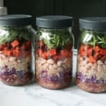 3 mason jars full of salad ingredients like sweet potato, white beans, cabbage, and arugula.
