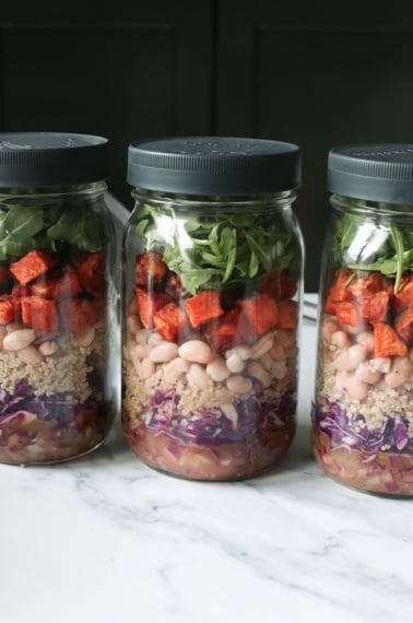 3 mason jars full of salad ingredients like sweet potato, white beans, cabbage, and arugula.