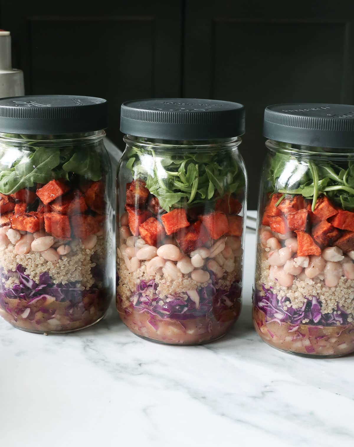 3 mason jars full of salad ingredients like sweet potato, white beans, cabbage, and arugula.