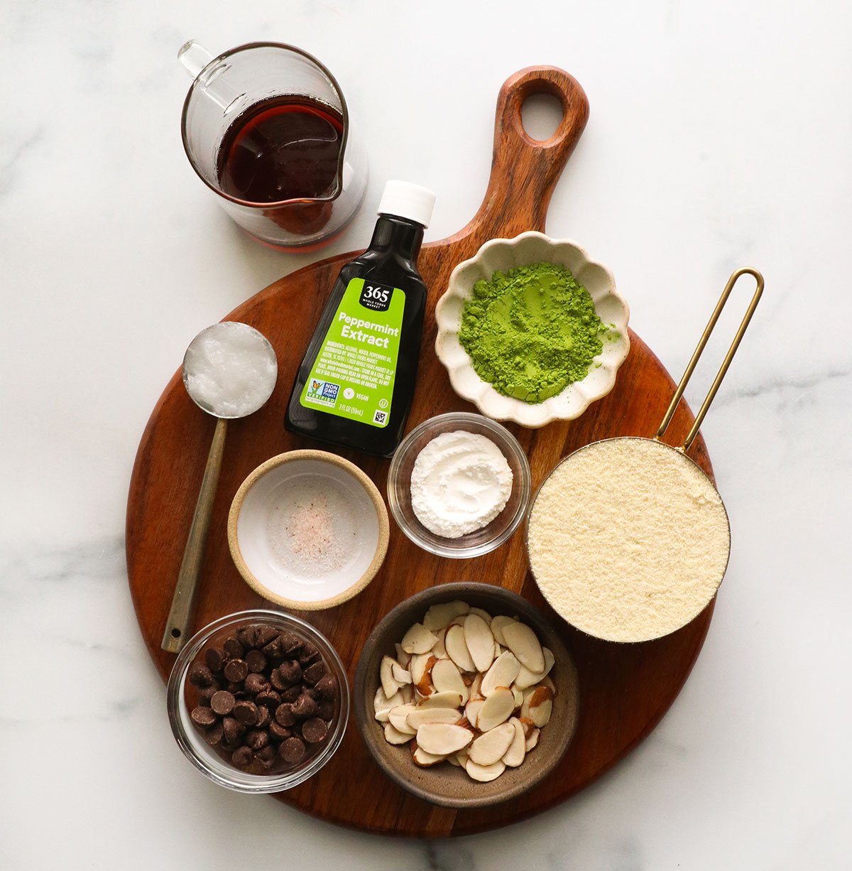 cups of almond flour, matcha, sliced almonds, chocolate chips, maple syrup, and coconut oil on a wooden board.