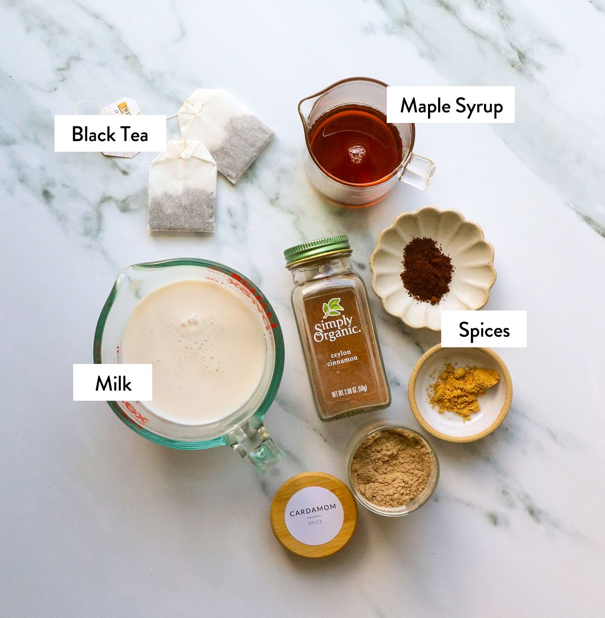Milk, tea bags, maple syrup and spices marked on a white surface.