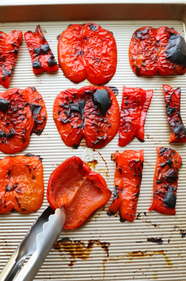 roasted red peppers on a baking sheet with one getting flipped over by a pair of tongs.