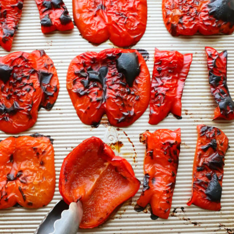 roasted red peppers on a baking sheet with one getting flipped over by a pair of tongs.