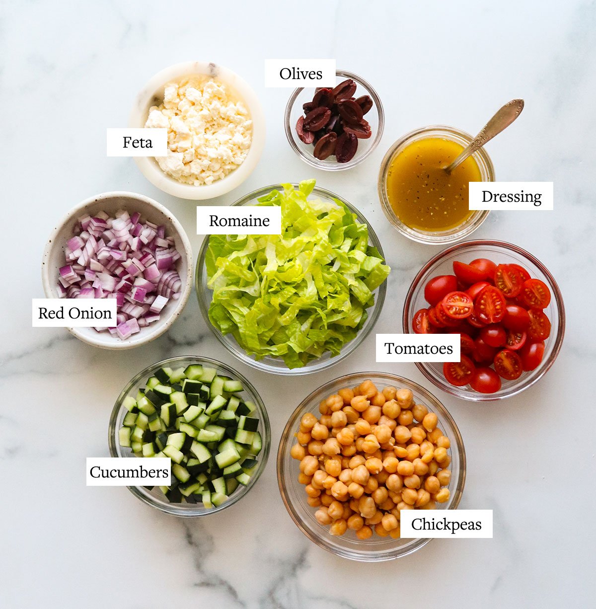 Bowls of ingredients labeled for mediterranean salad on a marble surface.