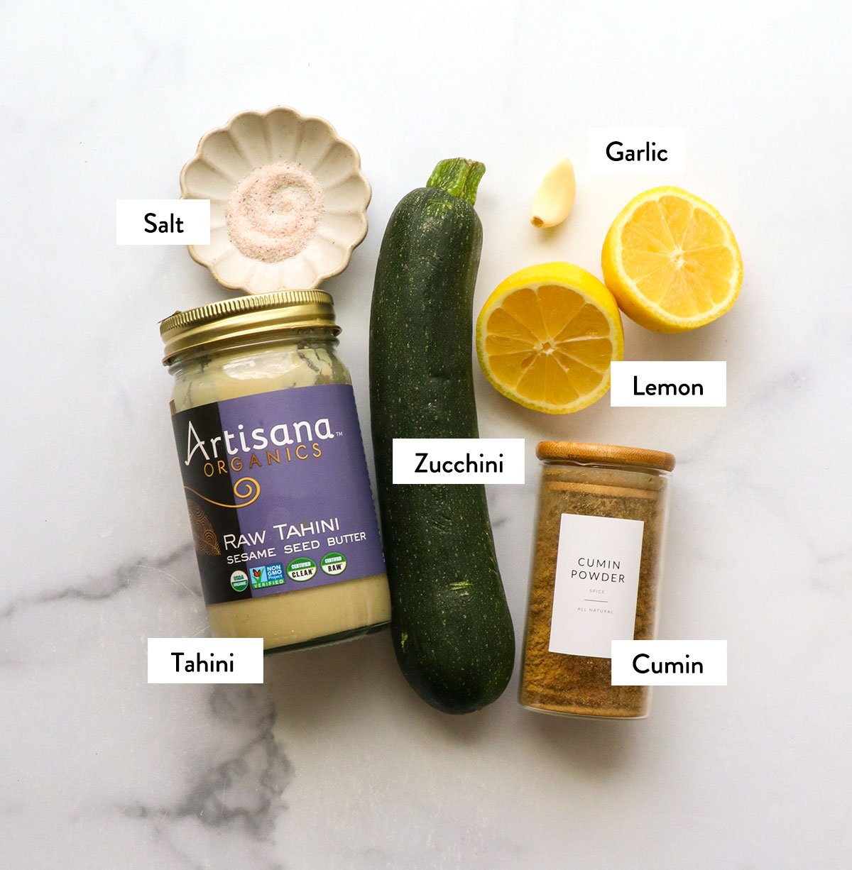 zucchini, tahini, lemon juice, and garlic labeled on a white surface. 