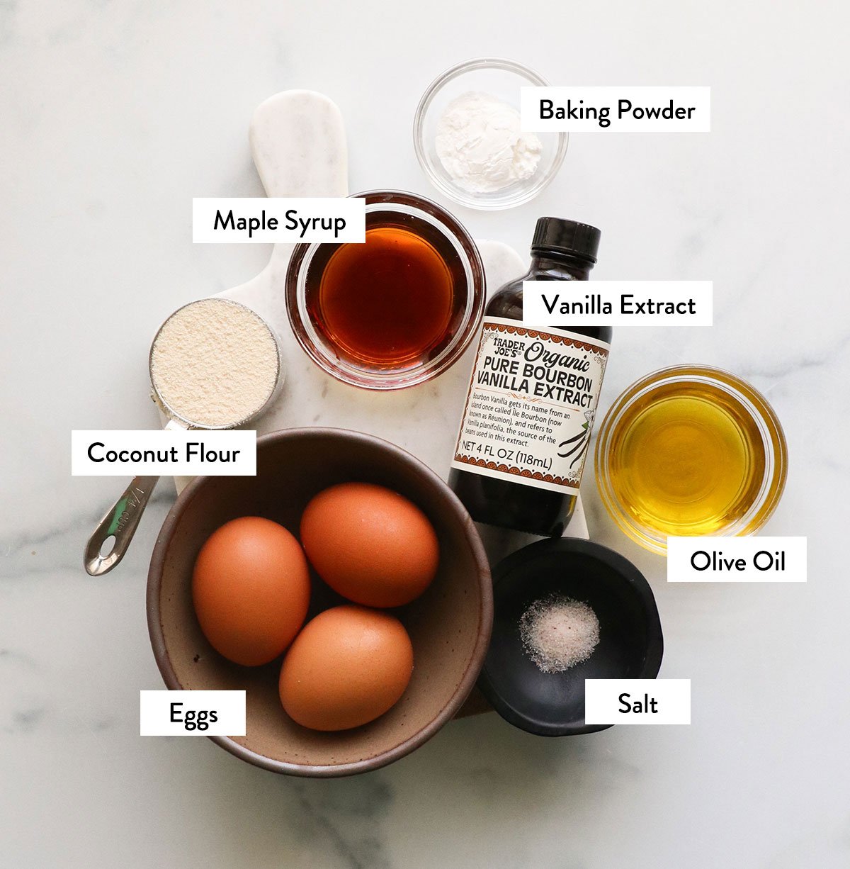 eggs, coconut flour, maple syrup, olive oil, and vanilla labeled on a white surface.