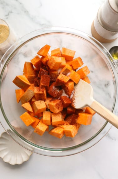 sweet potatoes seasoned in a glass bowl with a spatula.