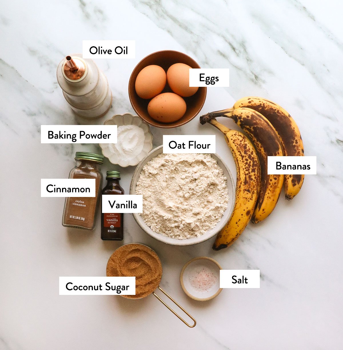 oat flour, bananas, eggs, oil, coconut sugar, and spices labeled on a white surface. 