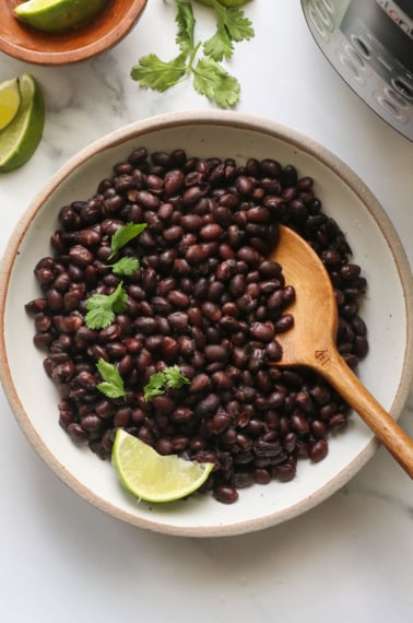 black beans in a white bowl with wooden spoon and a lime by the pressure cooker.