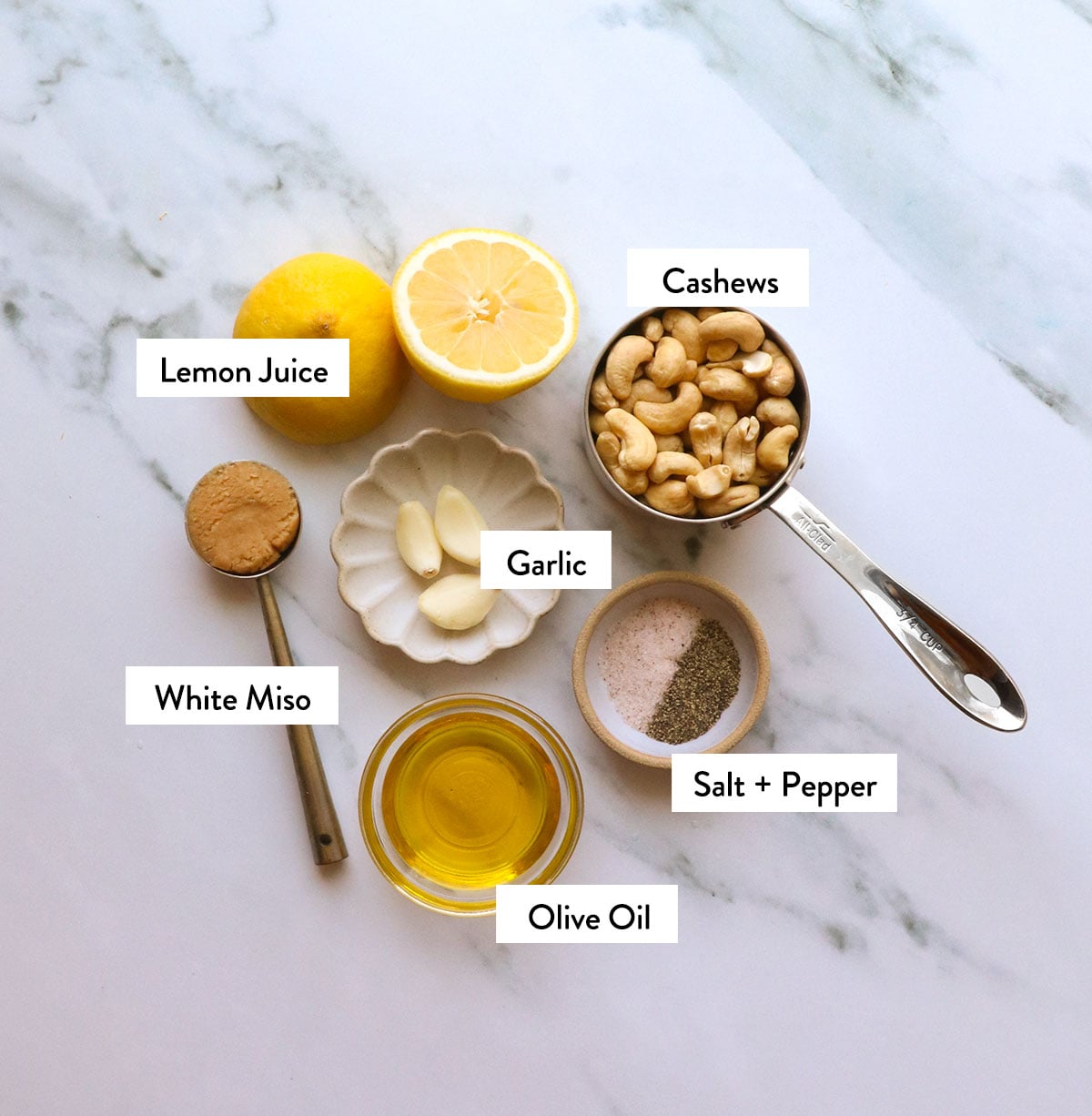 cashews, lemon, miso, olive oil, garlic, and spices labeled on a white surface.