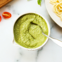 zucchini pesto in a white bowl next to a plate of pasta and cherry tomatoes.