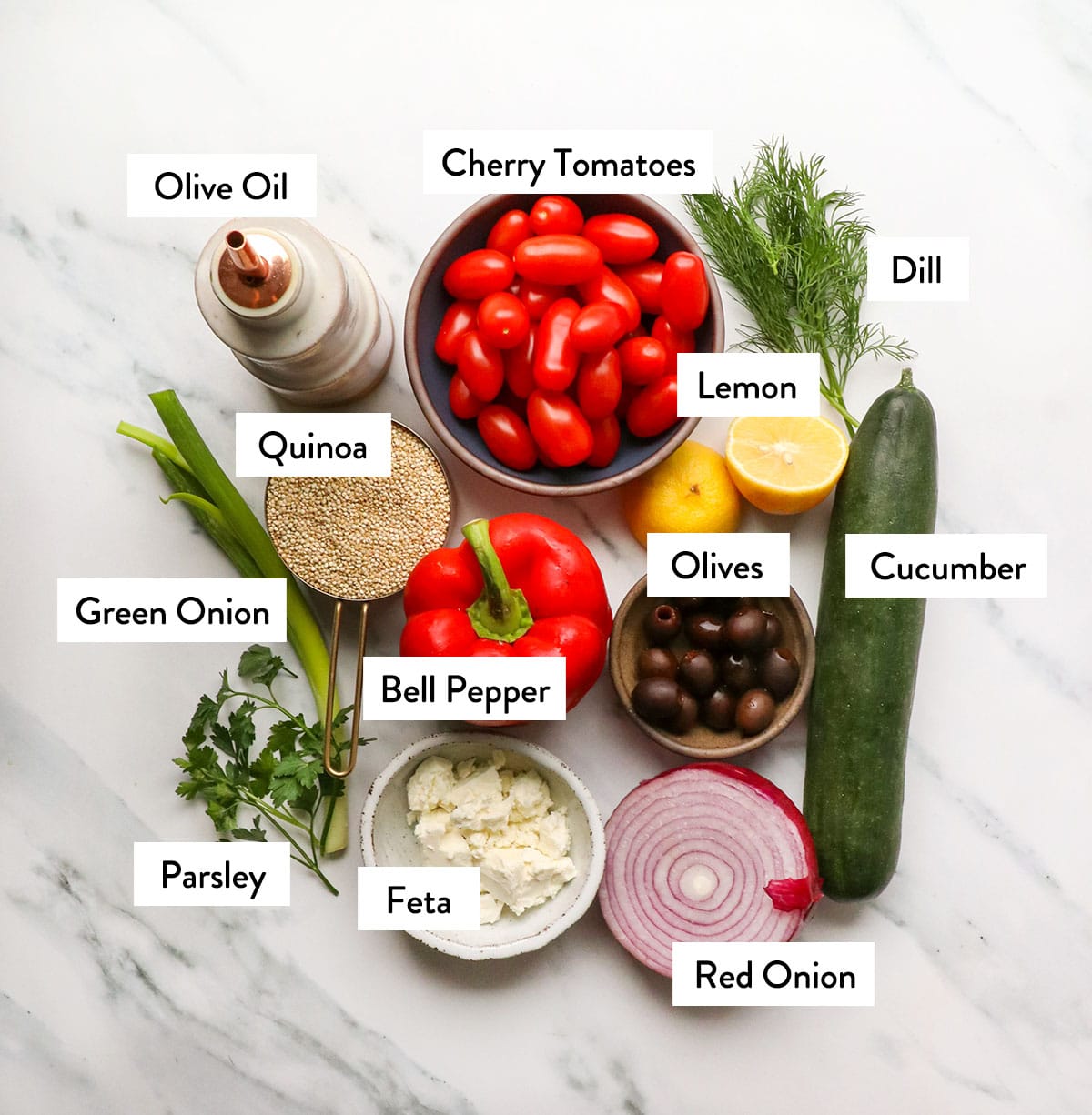 bowl of tomatoes, olive oil, quinoa, lemon, feta, cucumber, red onion, and parsley on a white surface.