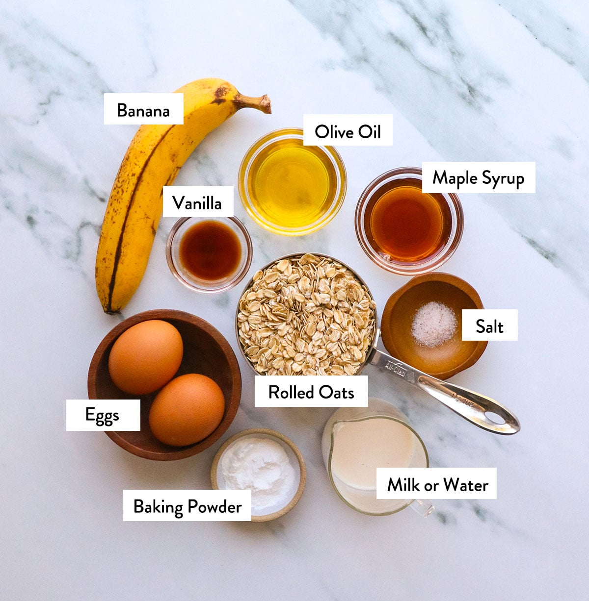 rolled oats, banana, maple syrup, baking powder, and eggs labeled on a white surface. 