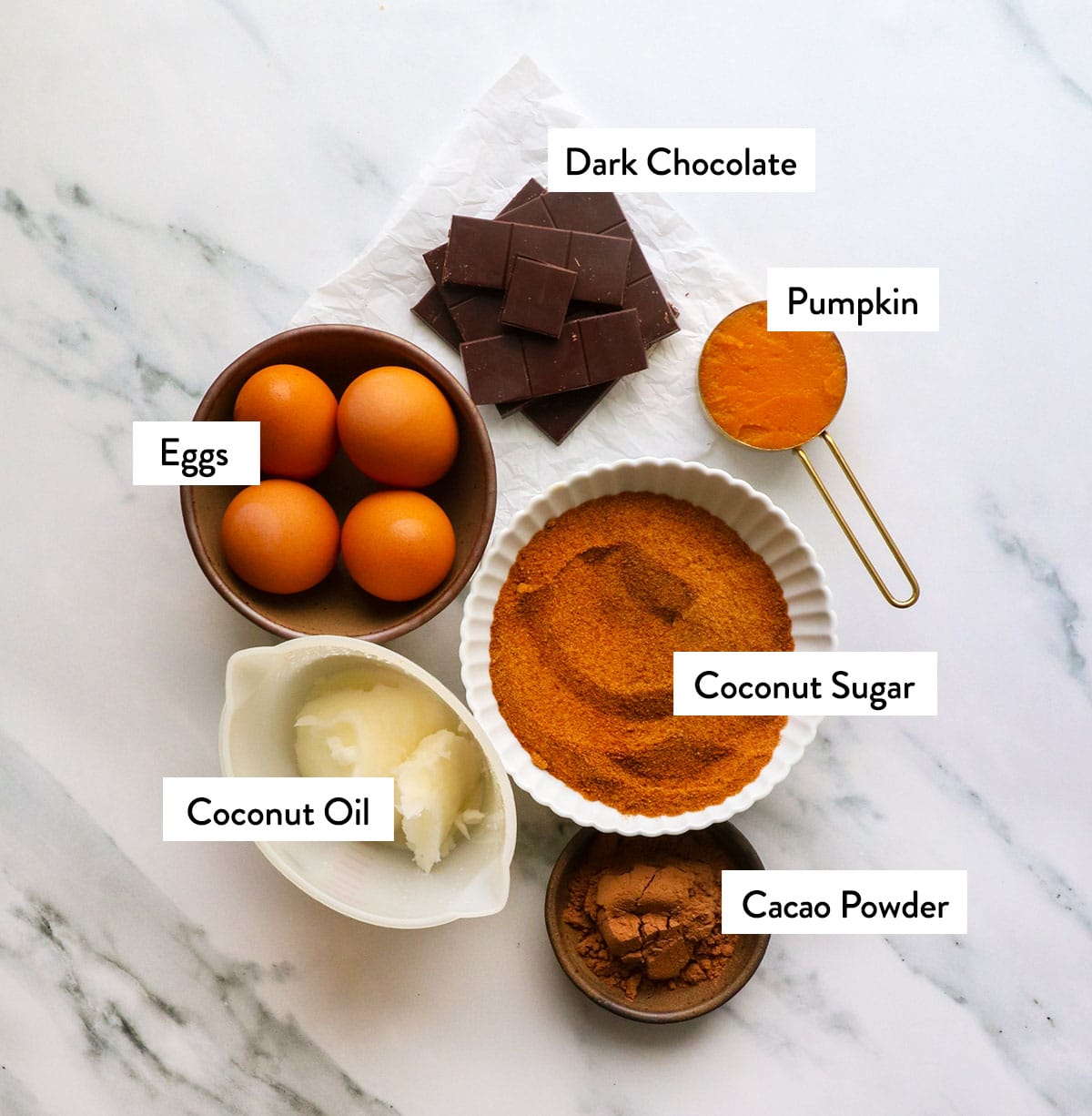 eggs, dark chocolate, coconut sugar, pumpkin, and coconut oil on a white surface.