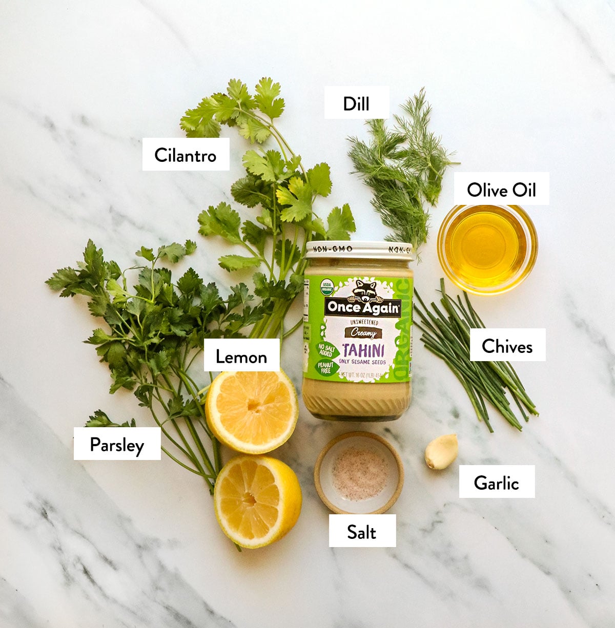fresh herbs, lemon, tahini, olive oil, garlic, and salt labeled on a white surface. 