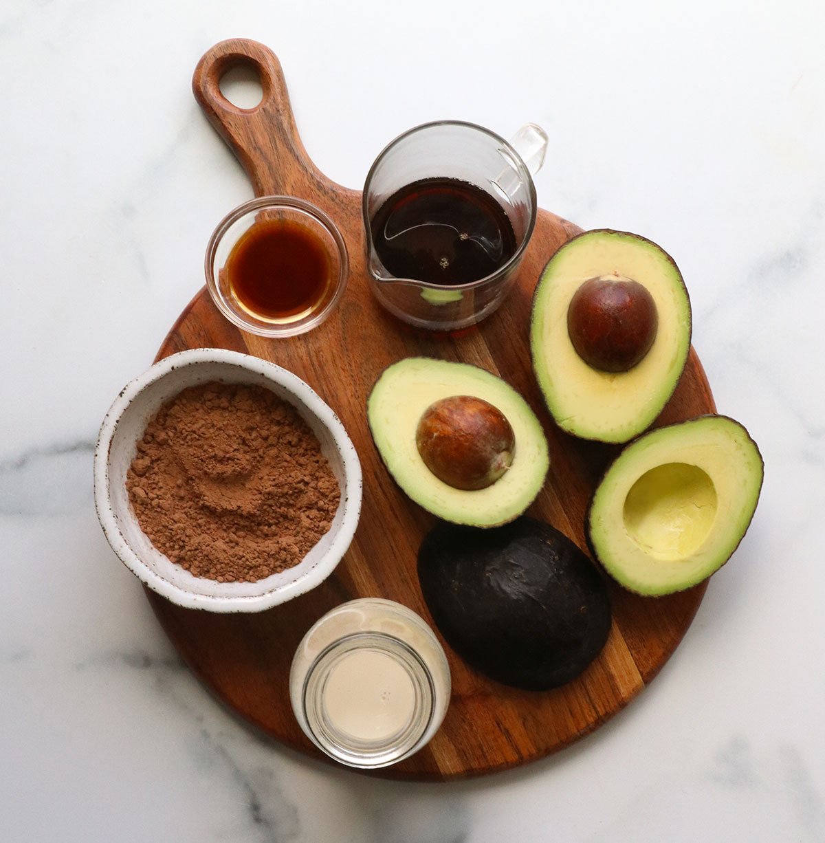 avocado, cocoa powder, maple syrup, milk, and vanilla extract arranged on a board.
