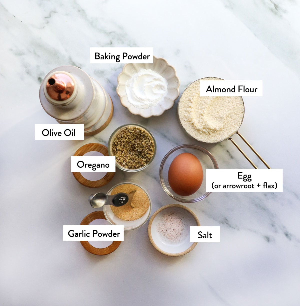 almond flour, baking powder, olive oil, spices, and an egg labeled on a white surface.