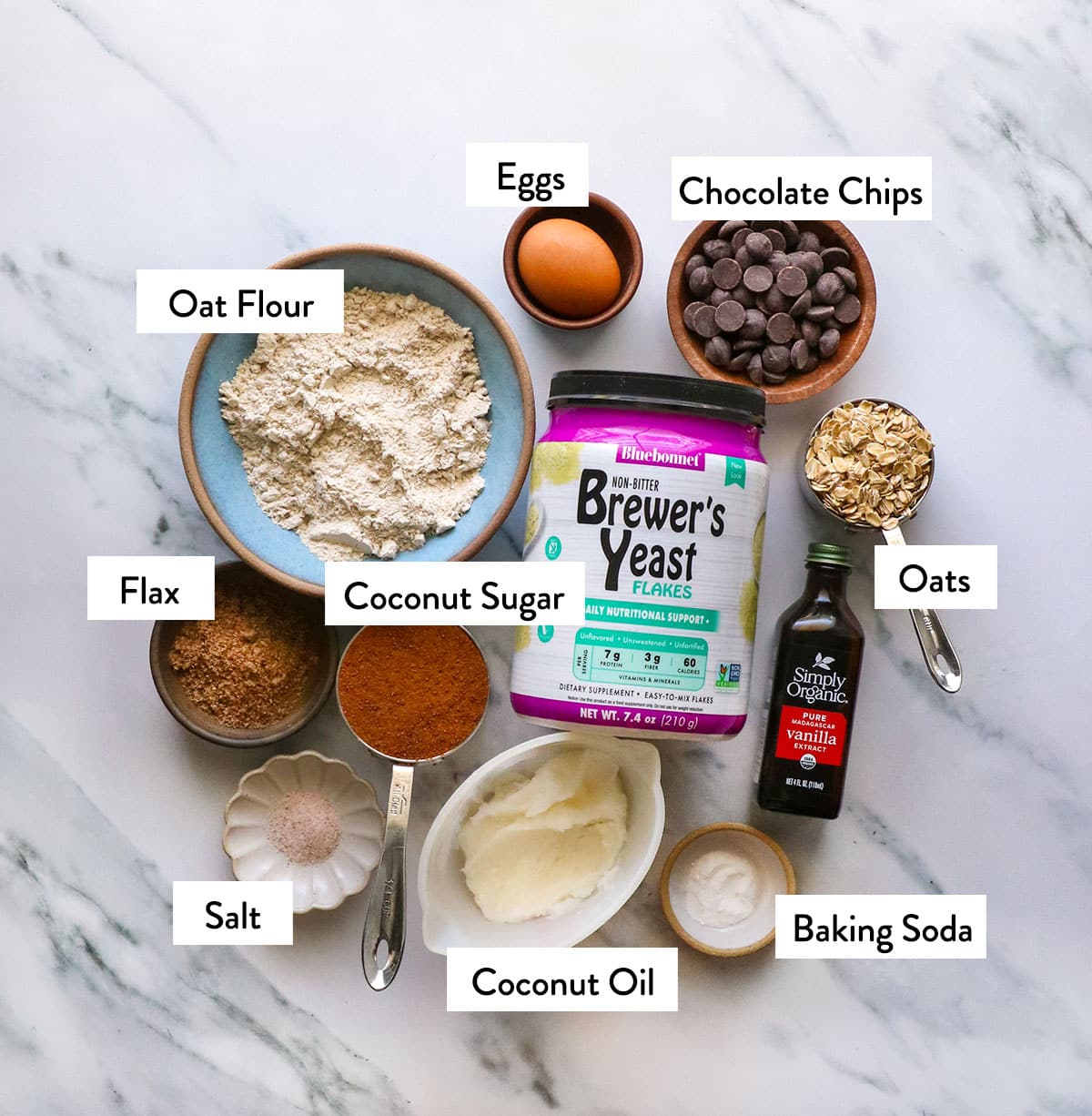 brewers yeast, oat flour, coconut oil, ground flax seed, chocolate chips, and oats in bowls on a white surface.