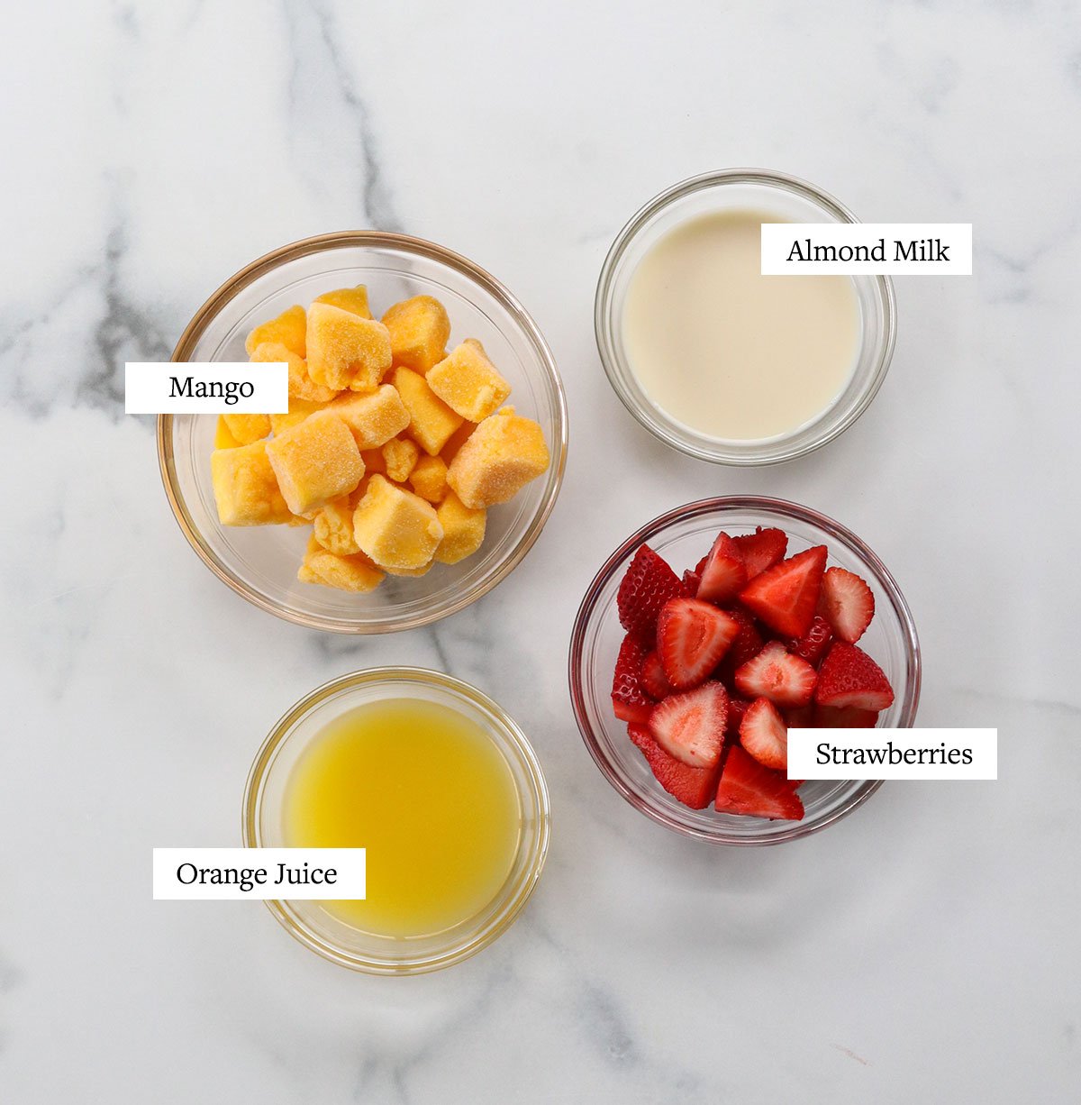 strawberry mango smoothie ingredients labeled in glass bowls on a white surface.