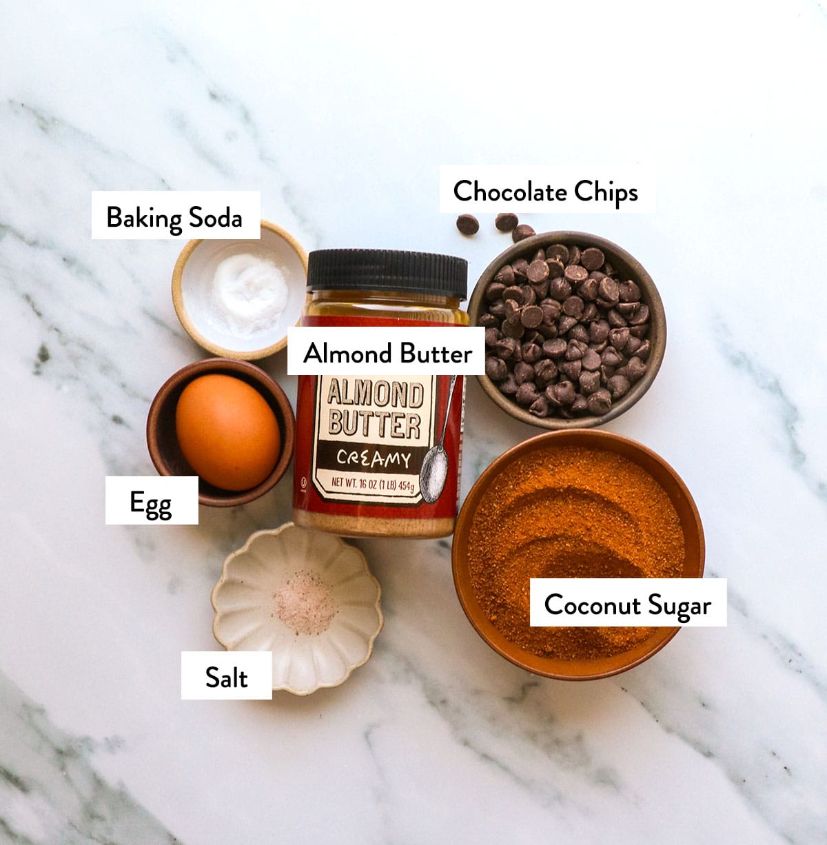 jar of almond butter, coconut sugar, egg, baking soda, and chocolate chips in bowls on a white surface.