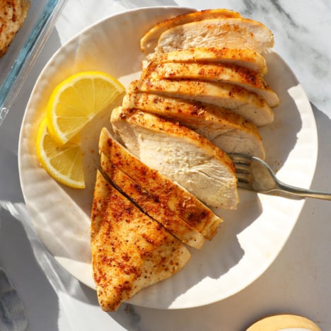 baked chicken breast sliced on a white plate with lemon slices nearby.