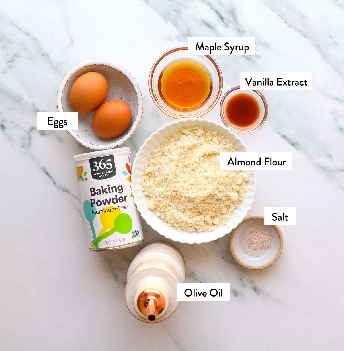 almond flour, maple syrup, eggs, vanilla, and oil labeled in bowls.