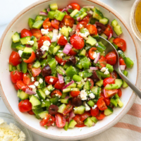 Greek salad in a white bowl with serving spoon and feta cheese on top.