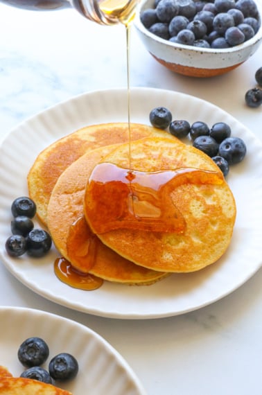 almond flour pancakes stacked on a white plate with maple syrup poured on top.