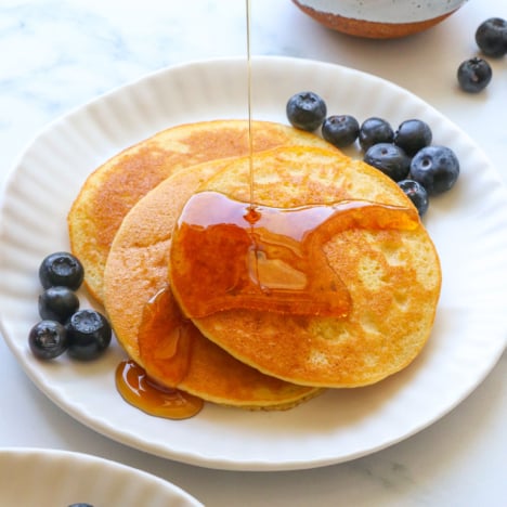 almond flour pancakes stacked on a white plate with maple syrup poured on top.
