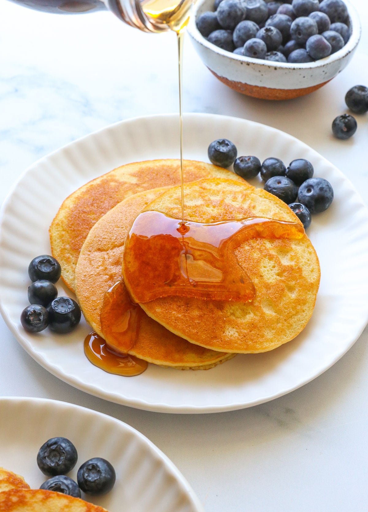 almond flour pancakes stacked on a white plate with maple syrup poured on top.