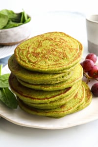 spinach pancakes stacked on a white plate by red grapes.