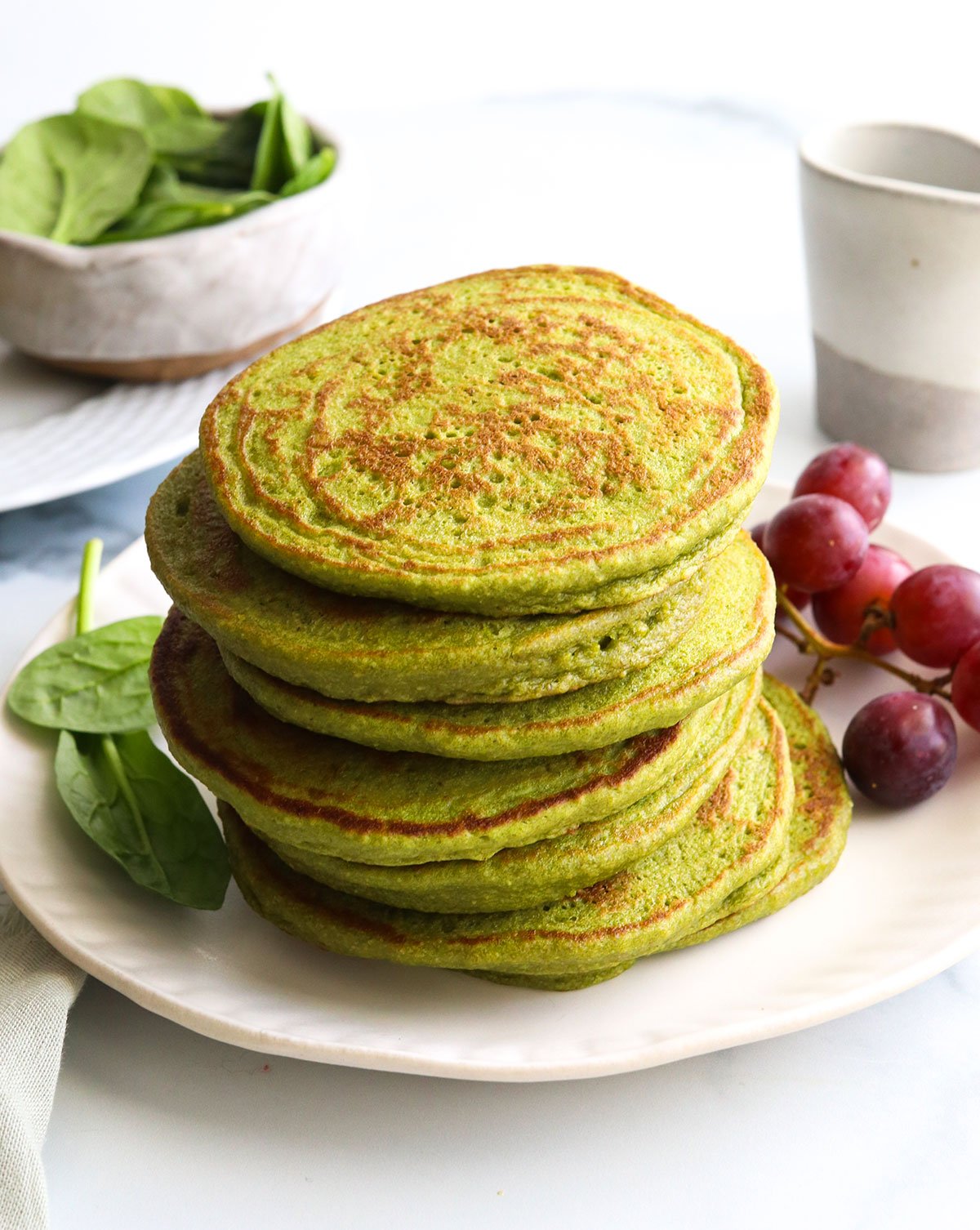 spinach pancakes stacked on a white plate by red grapes.