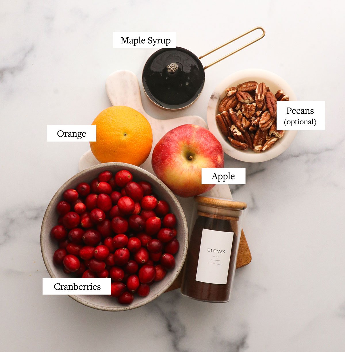 a bowl of cranberries, pecans, maple syrup, apple, orange, and cloves labeled on a white surface.