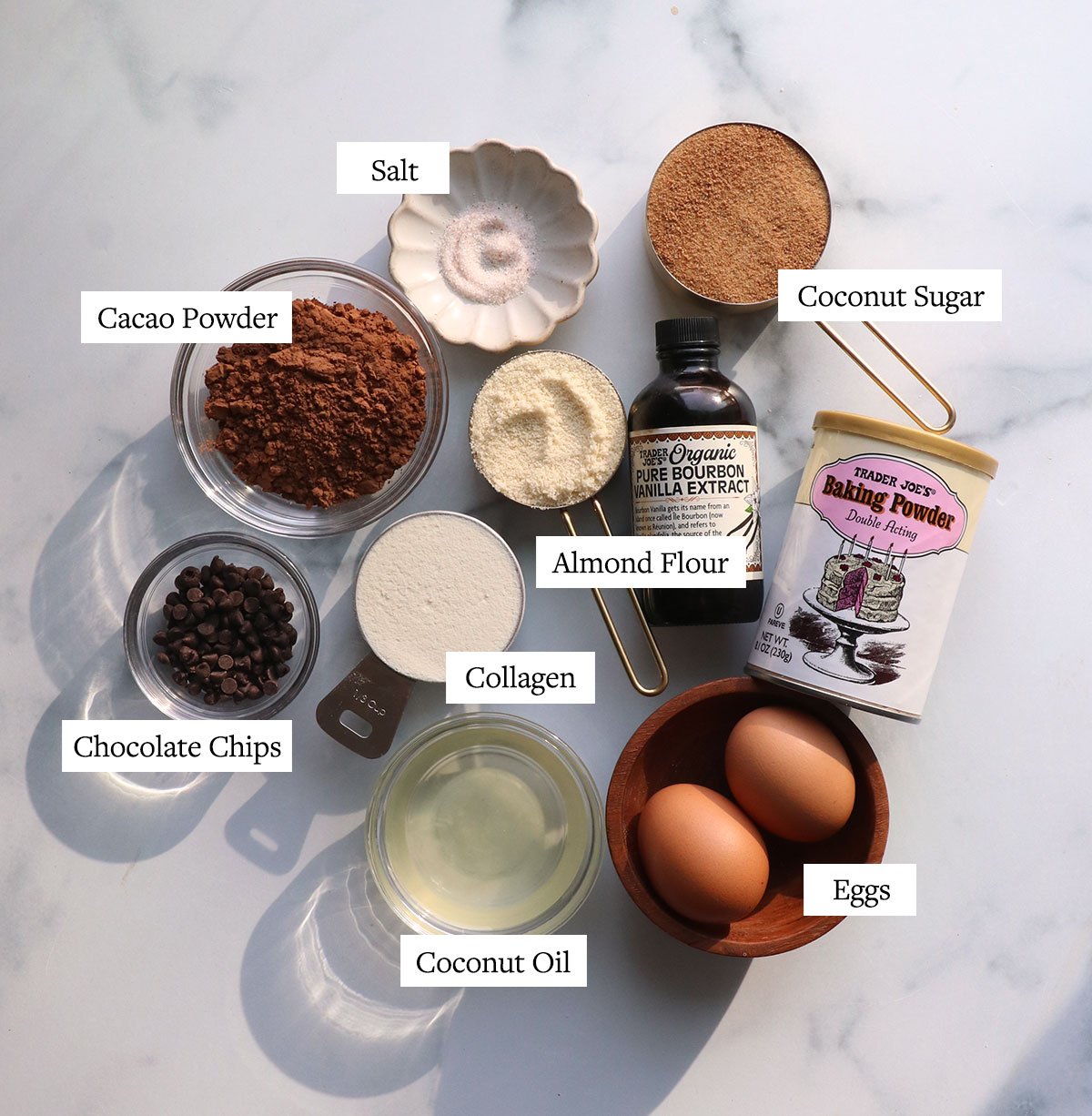 bowls of cacao powder, coconut sugar, collagen, almond flour and eggs labeled on a white surface.