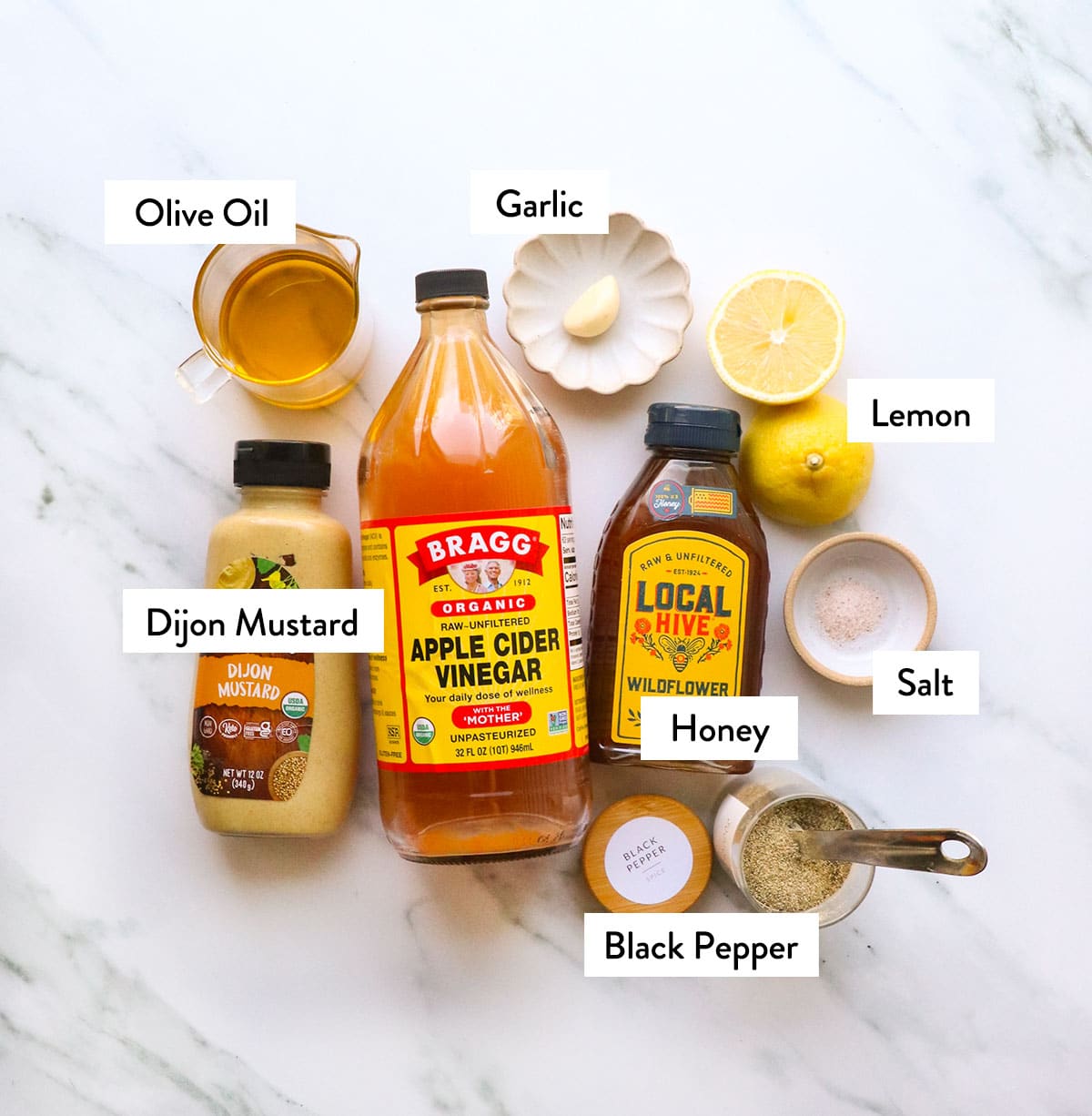 Bottles of apple cider vinegar, mustard, honey, olive oil, and a lemon on a white surface. 