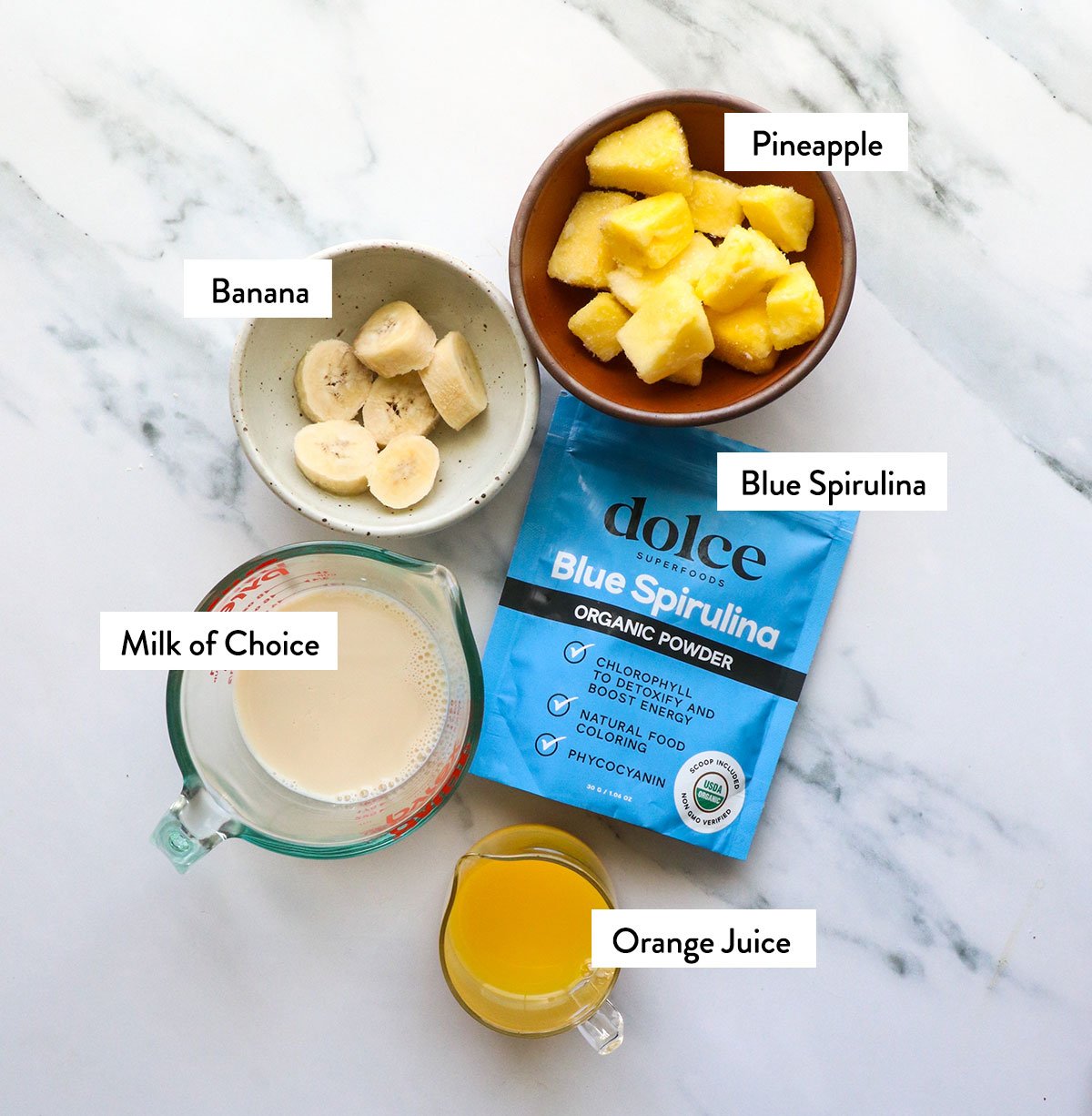 milk, frozen fruit, oj, and blue spirulina labeled on a white surface. 