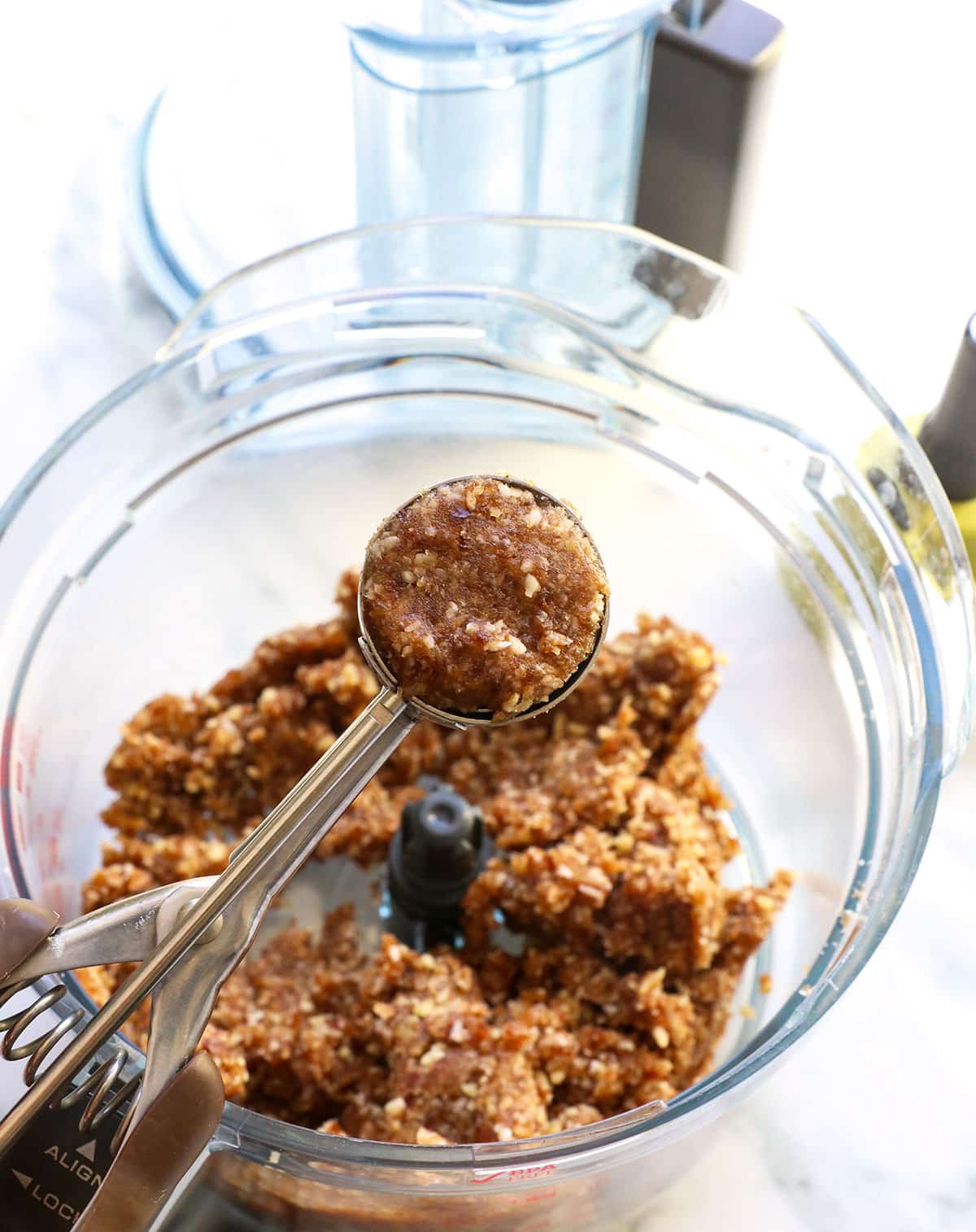 A cookie scoop lifting up some of the date ball mixture over the food processor.