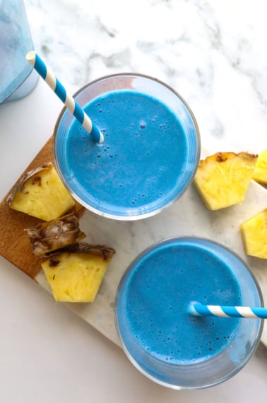 blue smoothie in two glasses with straws and pineapple nearby.