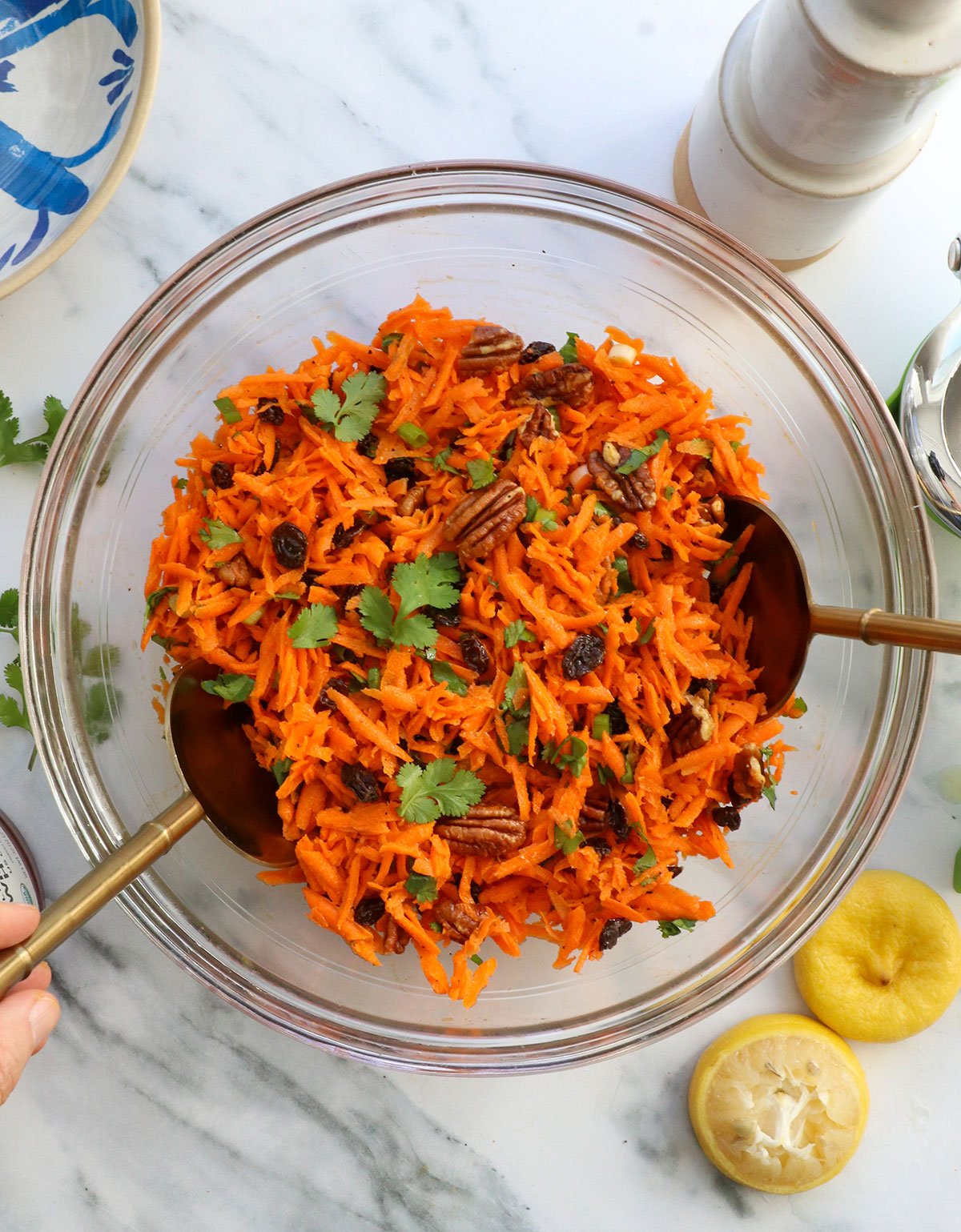 carrot salad with tongs in a glass bowl topped with pecans and raisins. 