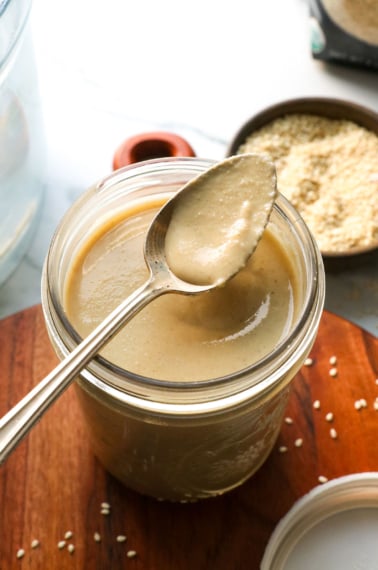 a spoon with homemade tahini sitting on a glass jar with more tahini.