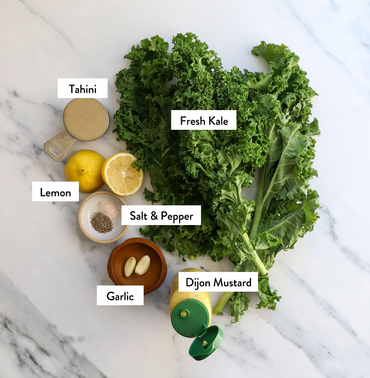 kale leaves, tahini, lemon, garlic, mustard, salt, and pepper labeled on a white surface. 