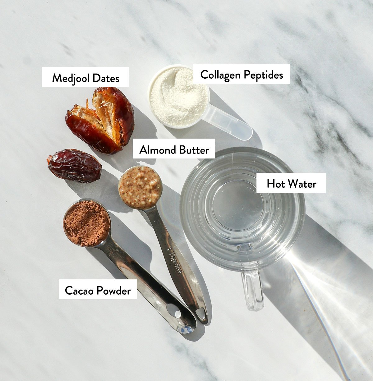 dates, almond butter, collagen, water, and cacao powder labeled on a white surface.