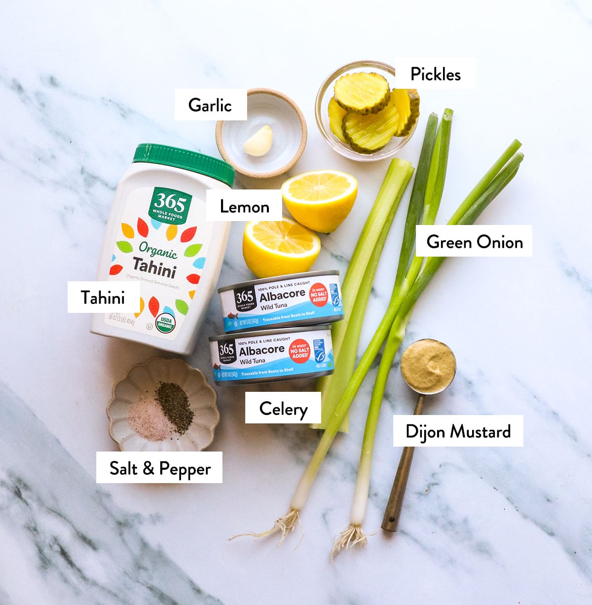 tahini, tuna, lemons, green onion, celery, and pickles labeled on a white surface.