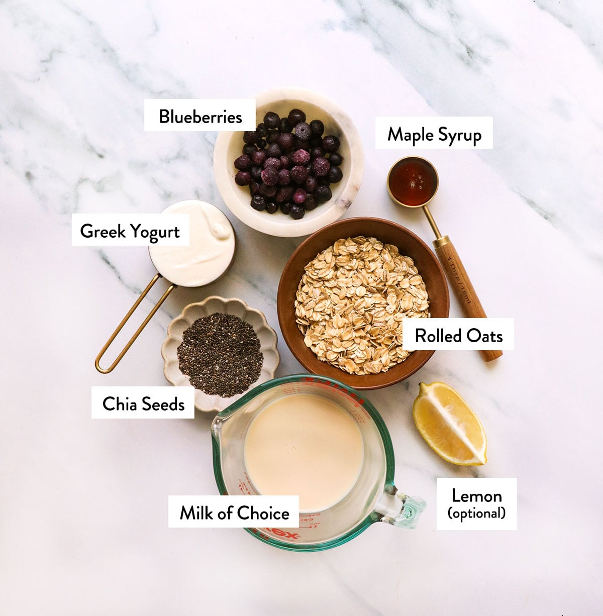 bowls of oats, blueberries, milk, chia seeds, yogurt, and milk on a white surface.